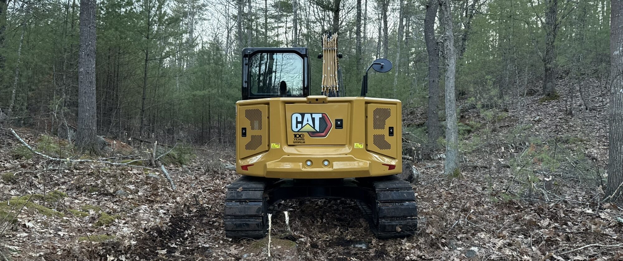 Cat excavator standing on a rough-cleared track through mixed Maine woodland, cutting the line for a private road.