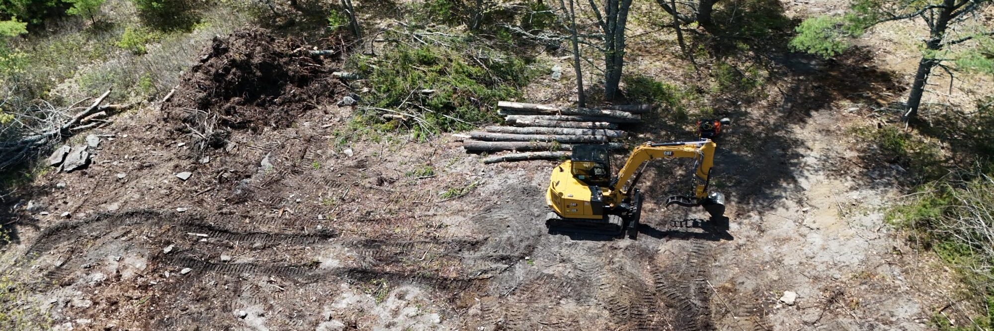 Aerial view of a partly cleared woodland lot in southern Maine, with a Cat excavator, stacked logs and a stump pile beside fresh machine tracks.
